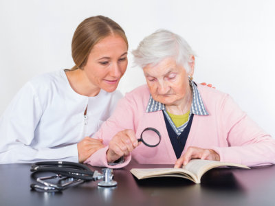 caregiver with elder woman reading a book with assistance of a magnifying glass