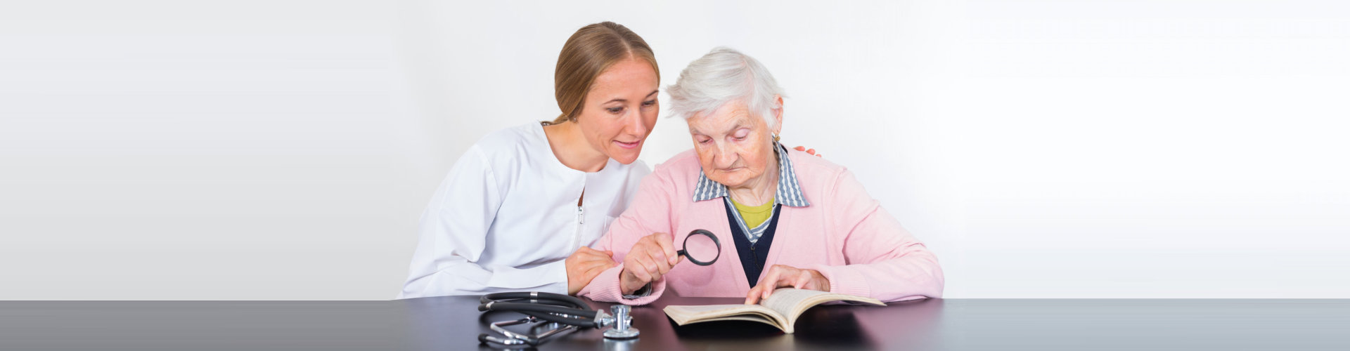 caregiver with elder woman reading a book with assistance of a magnifying glass